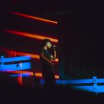 Trueno canta con un micrófono durante una actuación, iluminado por luces rojas y azules